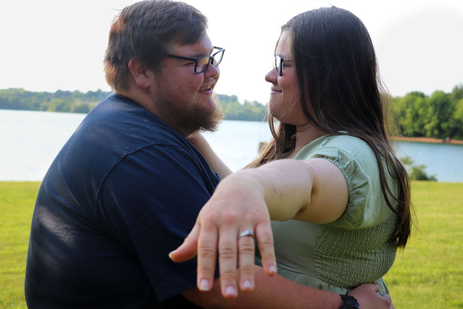 A joyful couple embraces and smiles at each other outdoors by a lake, showing off an engagement ring.
