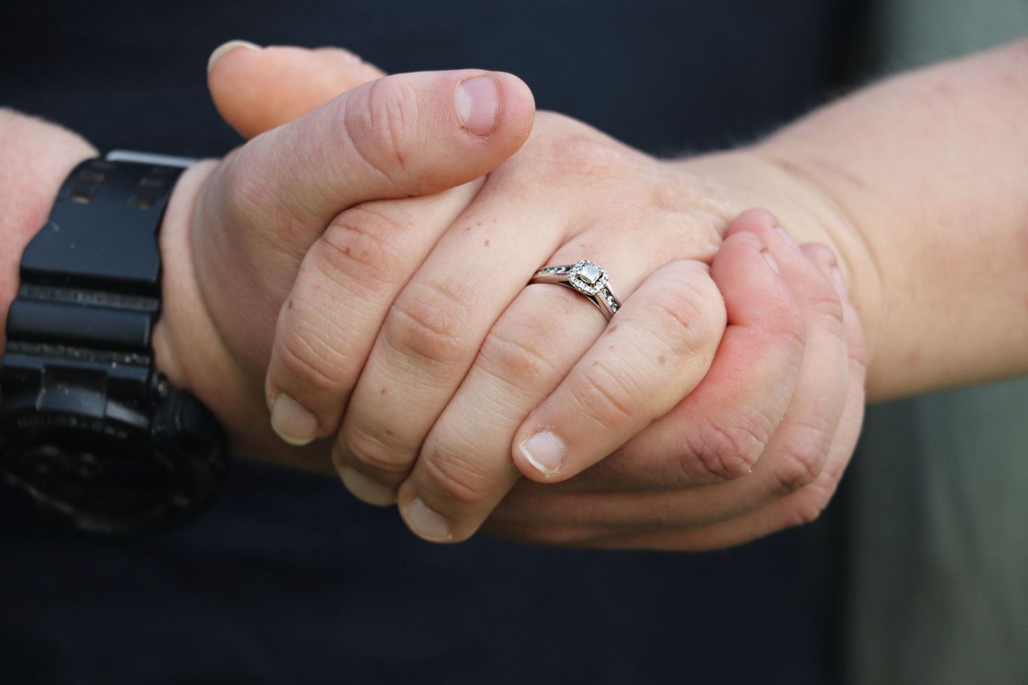 Close-up of a couple holding hands, showcasing a sparkling diamond engagement ring and a black watch.