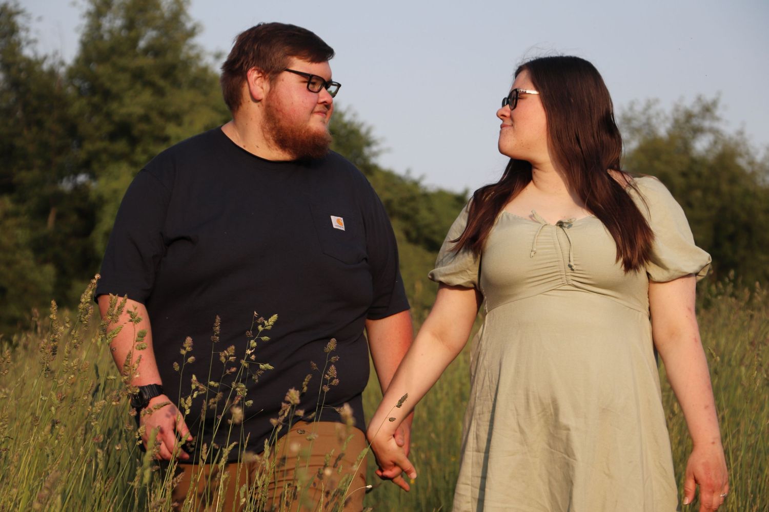 Couple holding hands walking through tall green grass field at golden hour.