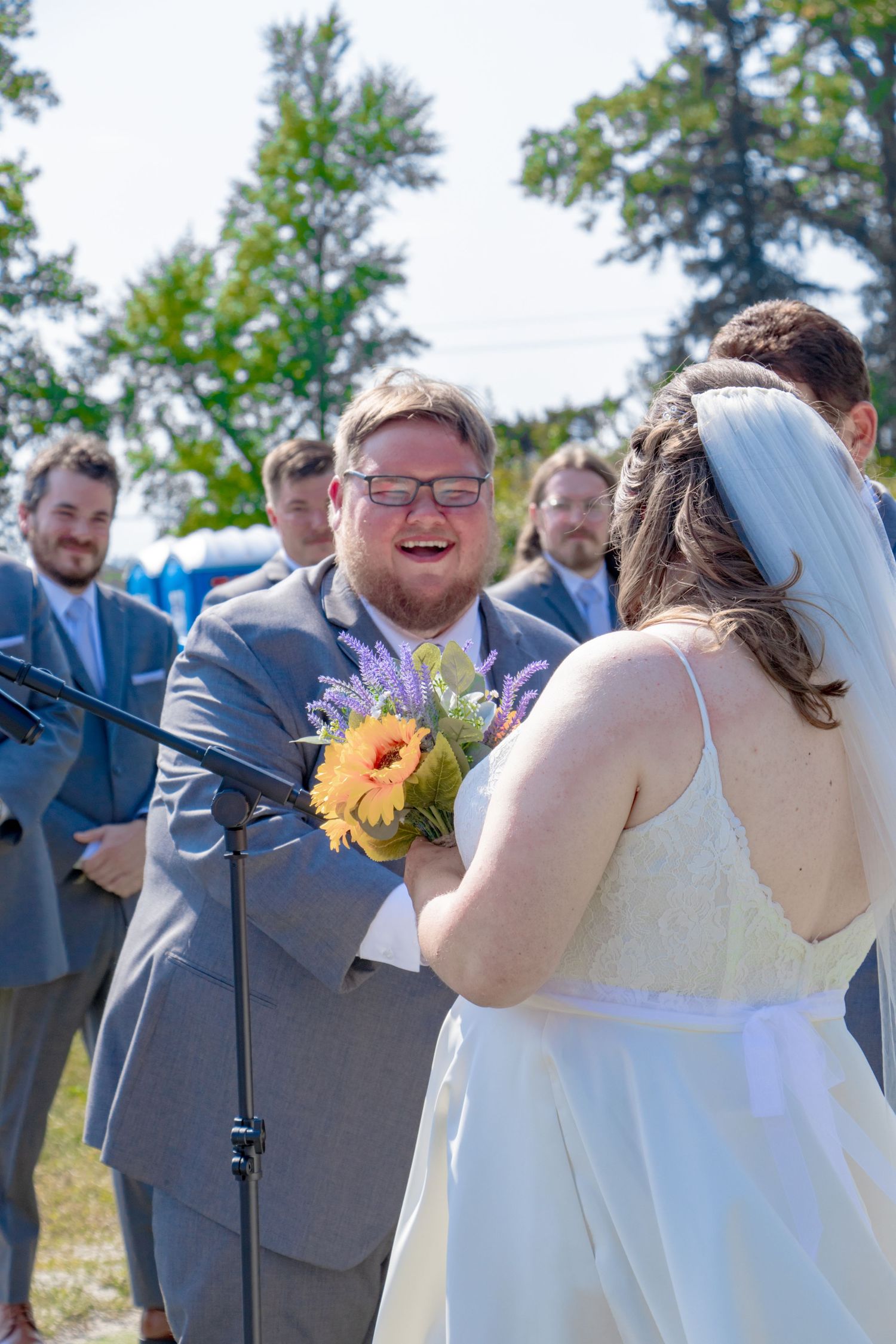 Groom laughing joyfully holding sunflower bouquet with bride at outdoor wedding.