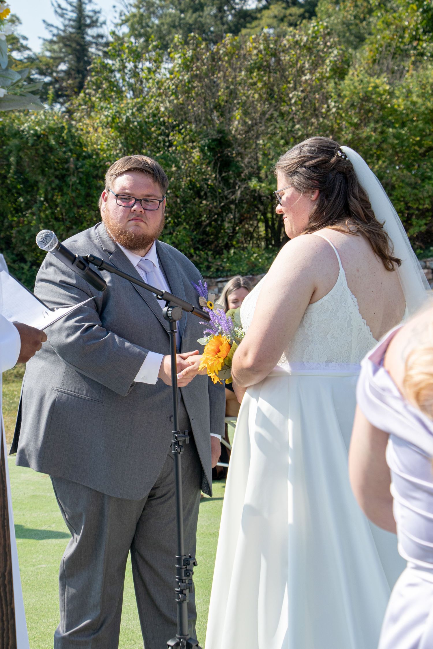 Groom in gray suit smiling at bride in white wedding gown during outdoor wedding ceremony.