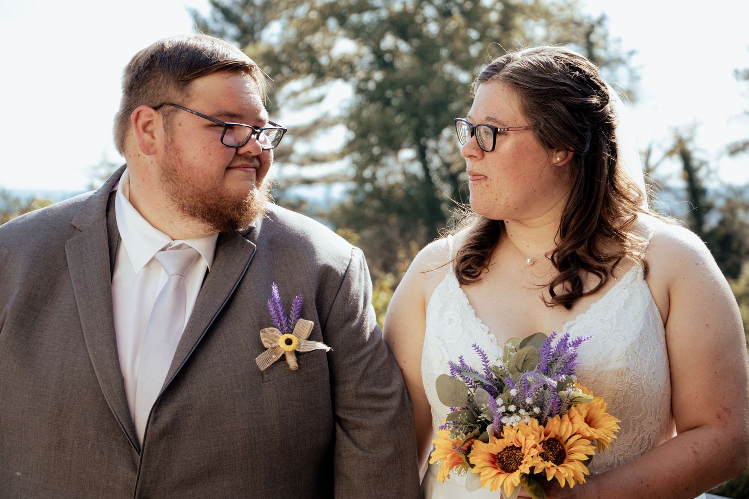 Newlyweds in gray suit and white gown holding sunflower bouquet smiling at each other.