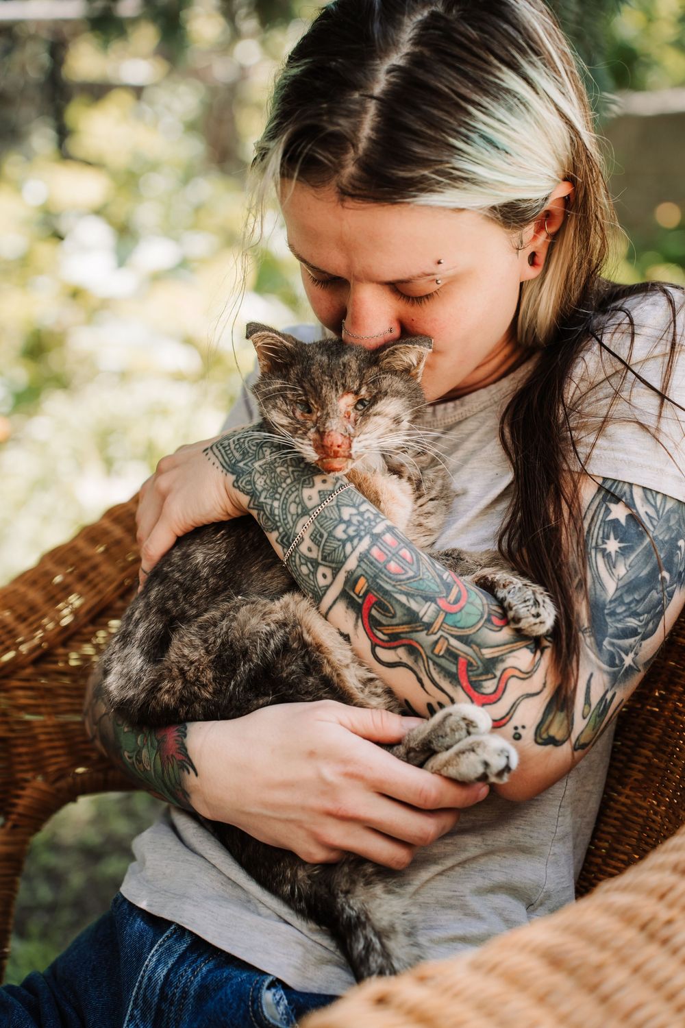 Tattooed woman tenderly holding and nuzzling a fluffy gray cat while sitting in a wicker chair outdoors.