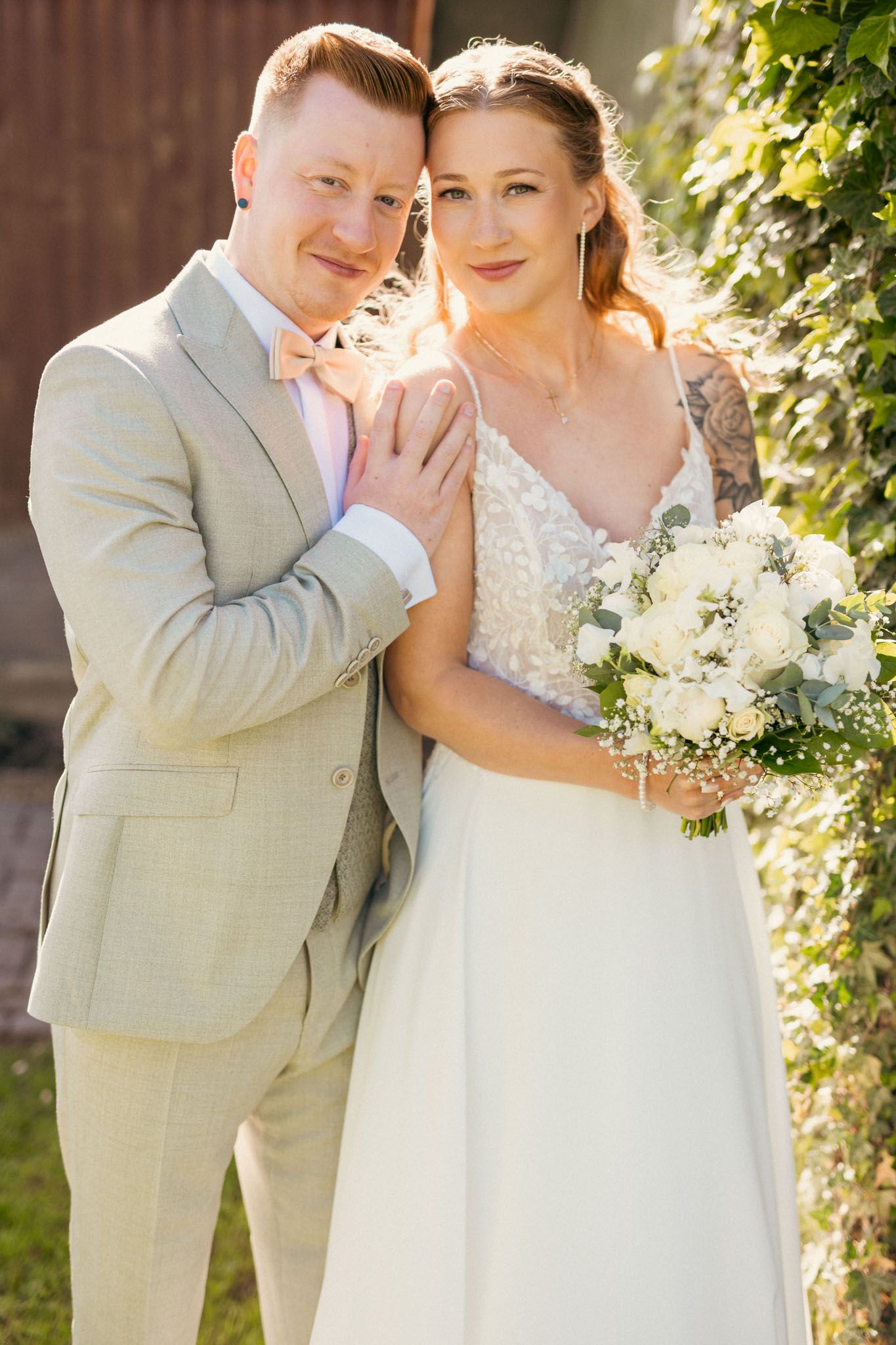 Bride and groom pose together outdoors, she holds white bouquet in wedding dress, he wears light gray suit with pink bow tie.