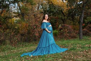 A flowing teal dress spreads elegantly across autumn leaves in a woodland setting during a photoshoot.