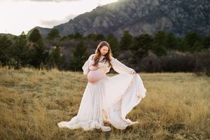 Pregnant woman in flowing white dress poses in golden field with mountain backdrop during sunset maternity photo session.