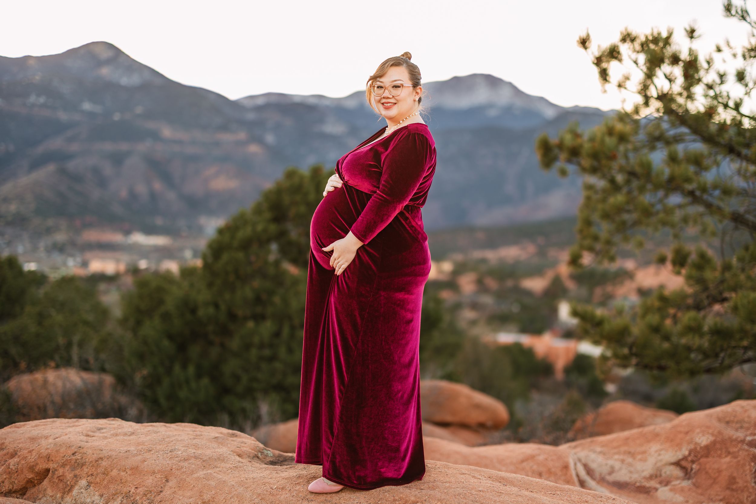 A person in a burgundy velvet dress poses gracefully against a backdrop of scenic mountain landscapes at sunset.