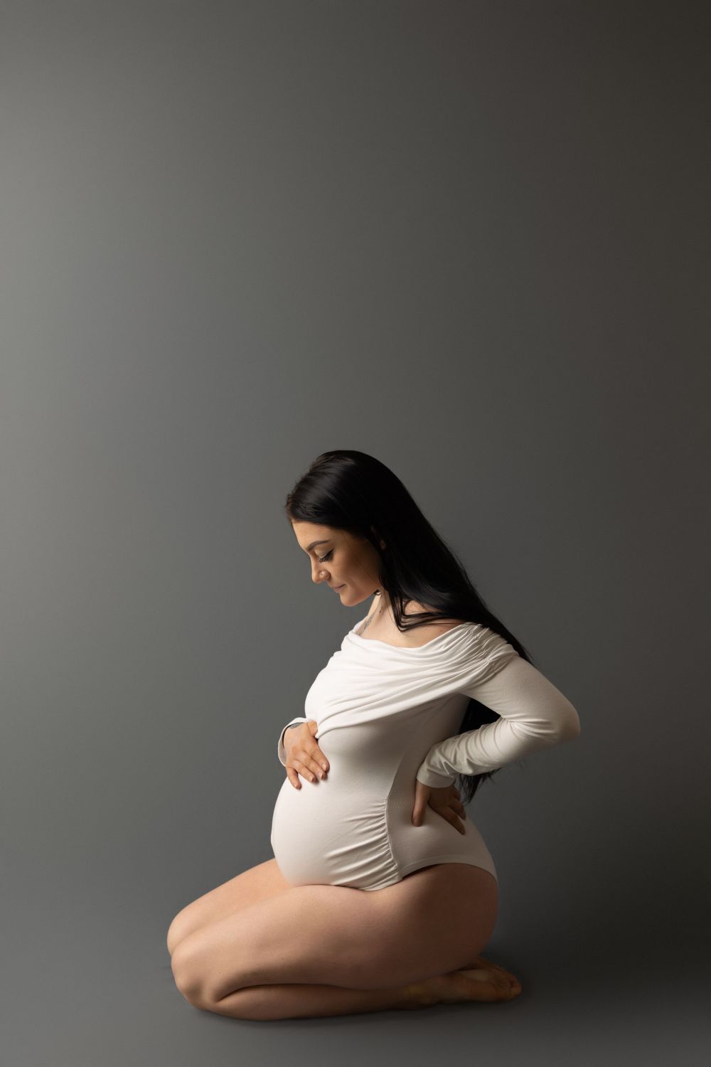 An artistic maternity pose in white bodysuit against a grey studio background.