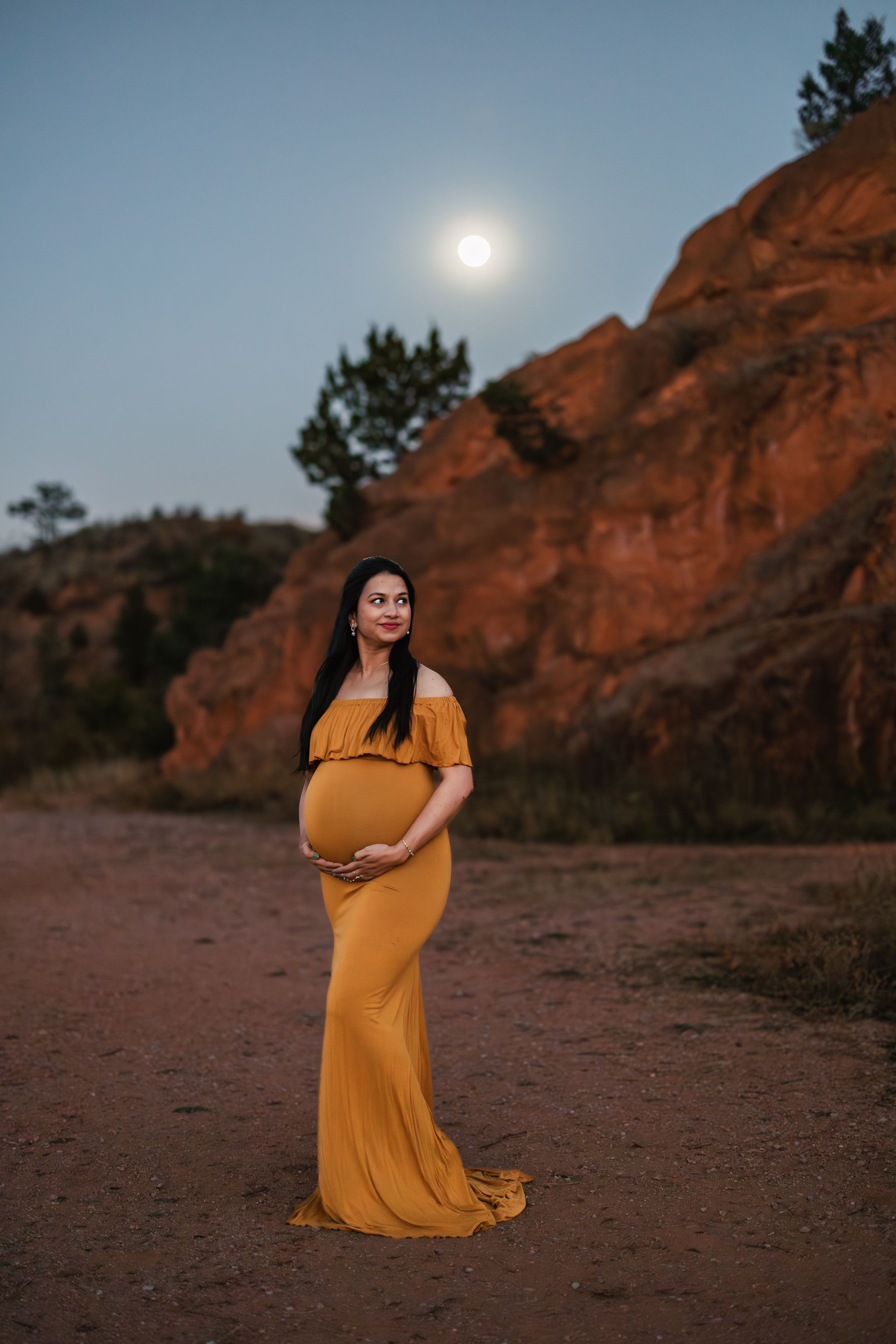 A woman in a flowing mustard yellow dress poses gracefully against red rock formations at sunset.