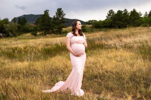 Stunning maternity portrait in a flowing pink gown against golden meadow and mountain backdrop at sunset.