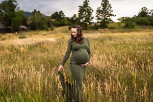 Expecting mother in olive green dress standing peacefully in tall grass field at dusk.