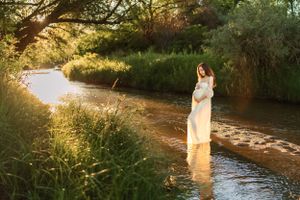 Dreamy maternity portrait in white dress standing in flowing stream during golden hour.