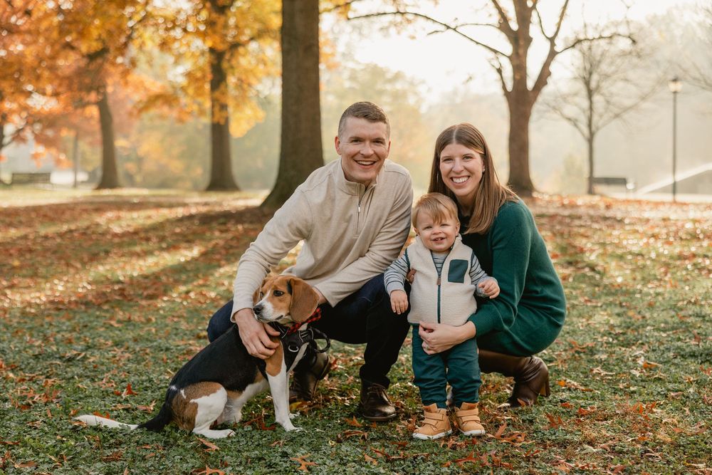 A family and their beagle dog pose together on fallen autumn leaves in a park during golden hour.