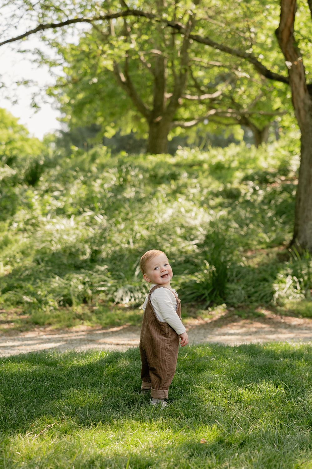 Happy toddler in brown overalls standing on green grass in a sunny park with lush trees.