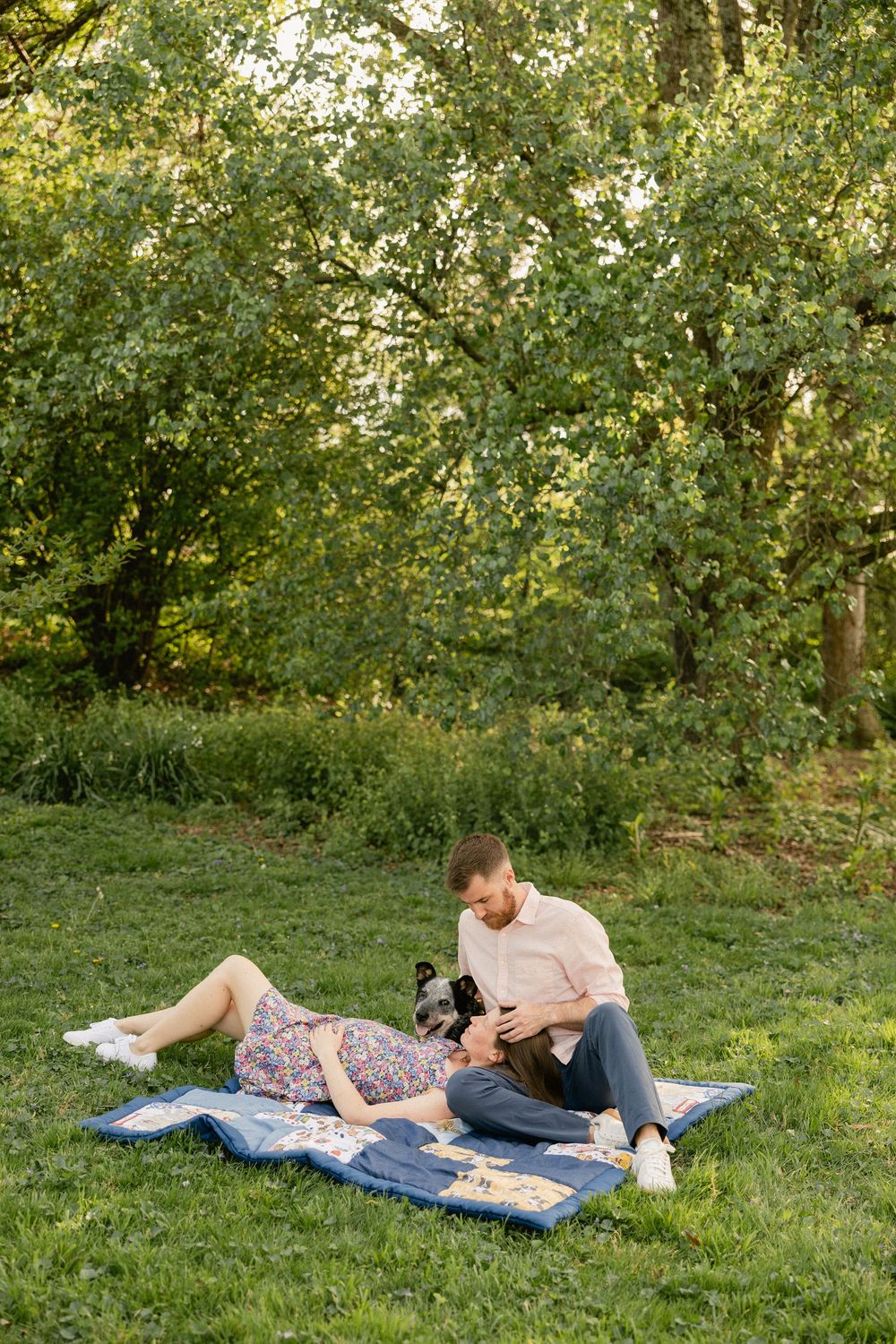 Couple relaxing on a patchwork quilt in a lush green park, with a small dog, enjoying a peaceful outdoor picnic.
