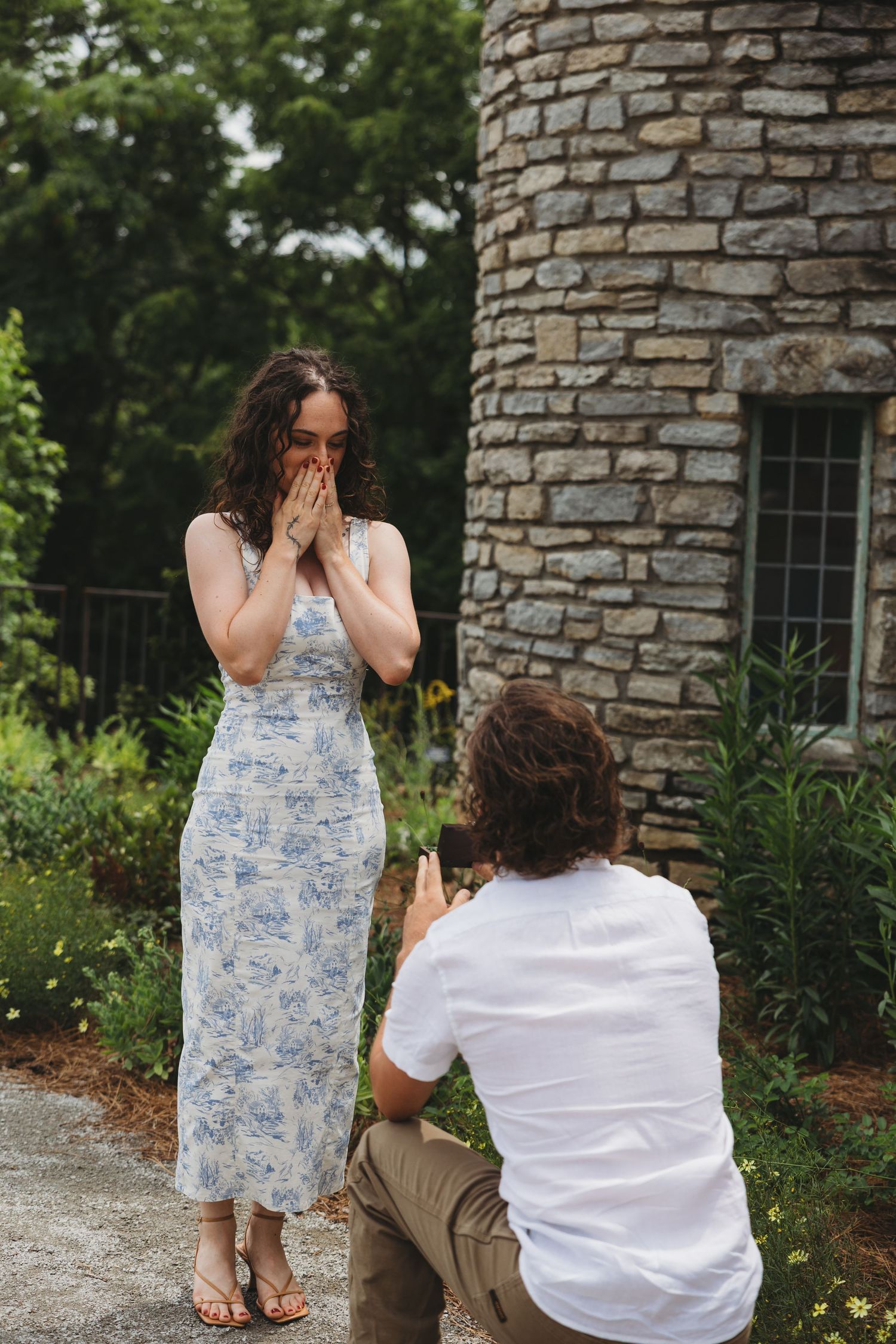 Man proposes on one knee to surprised woman in floral dress near stone building in lush garden setting.