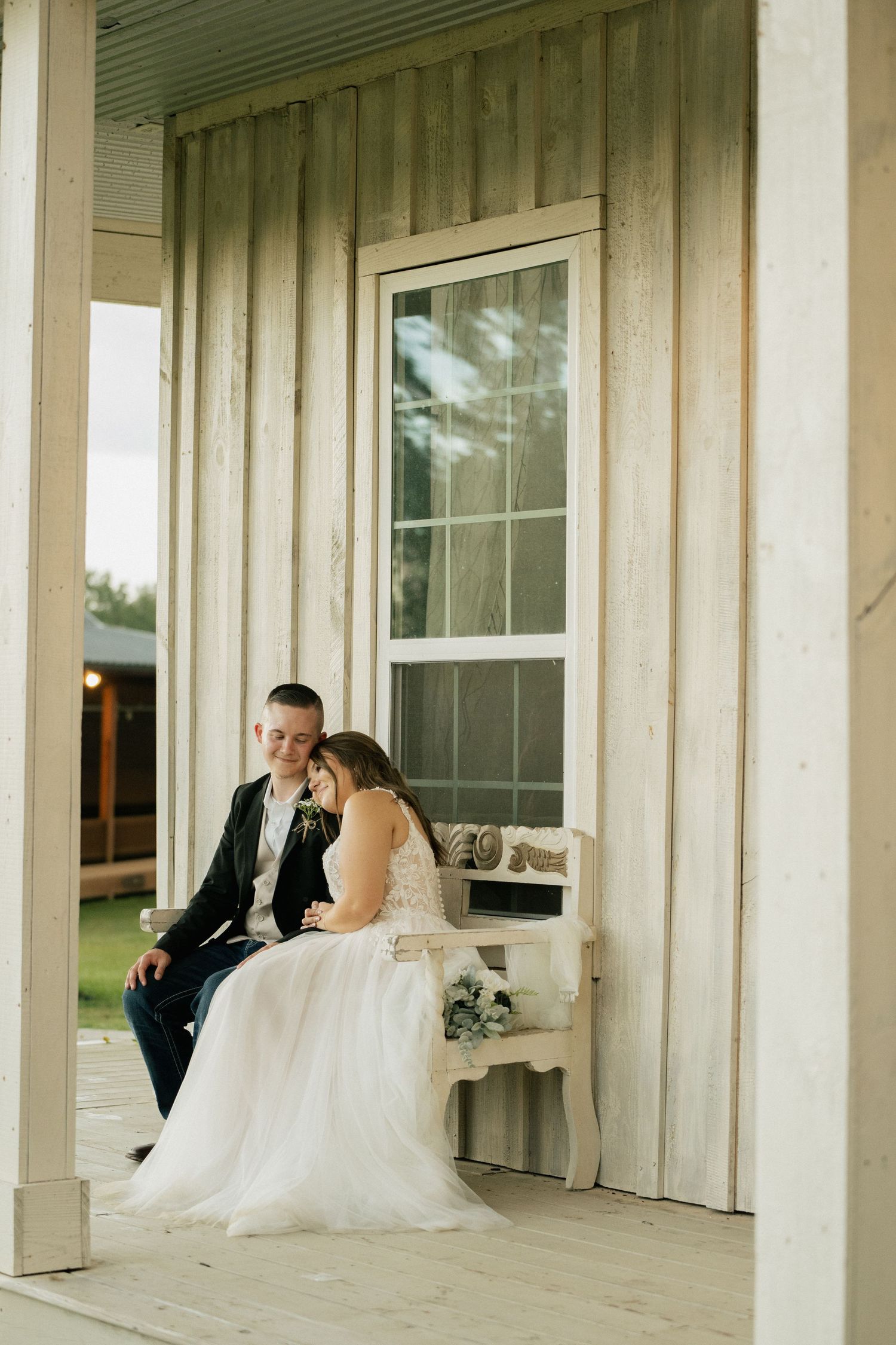A couple sits together on a rustic porch bench in wedding attire at dusk.