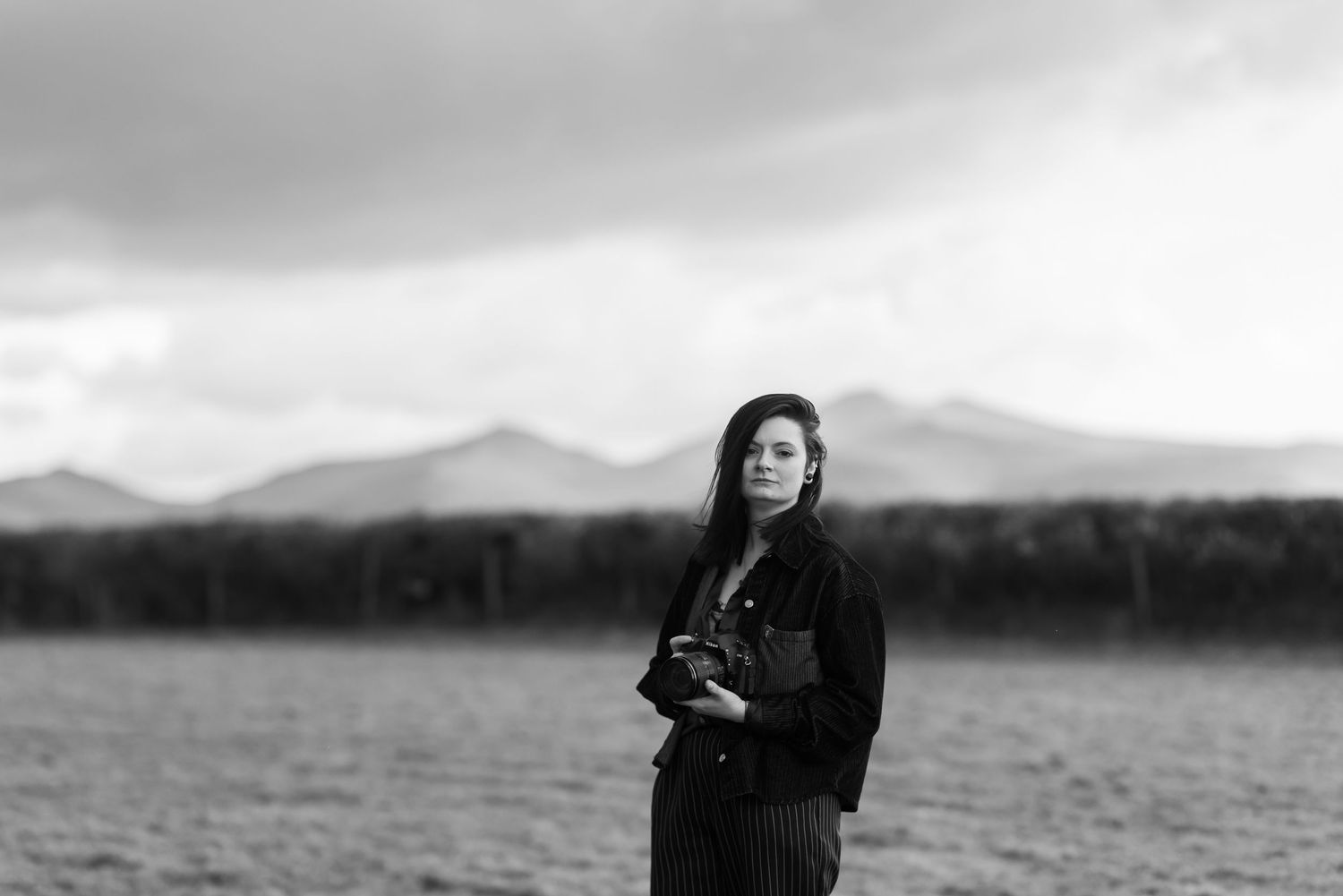 Black and white portrait of someone in plaid pants and boots posing with camera against brick wall.