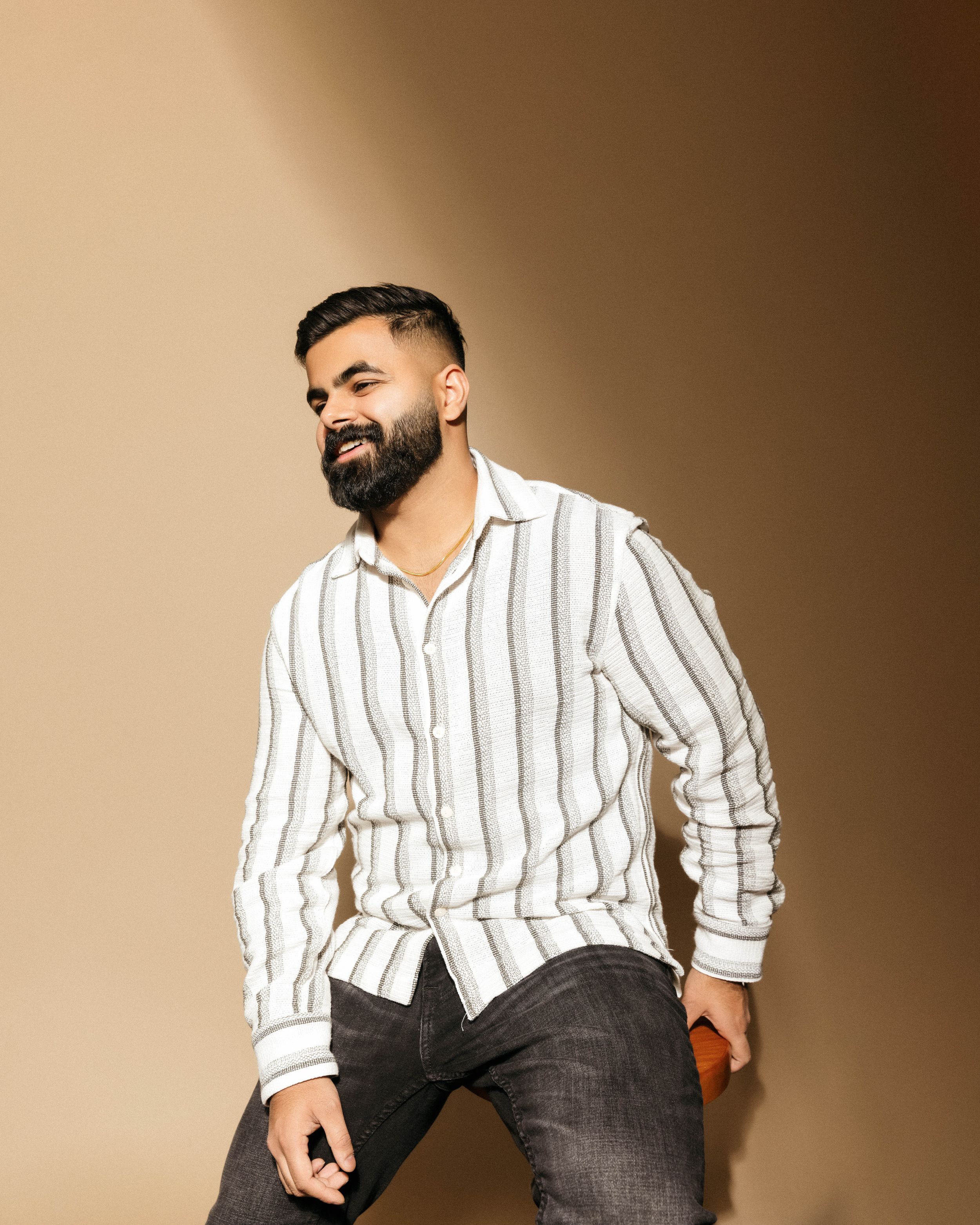 A man in a black jacket and pants poses casually while sitting on a wooden block against a gray background.