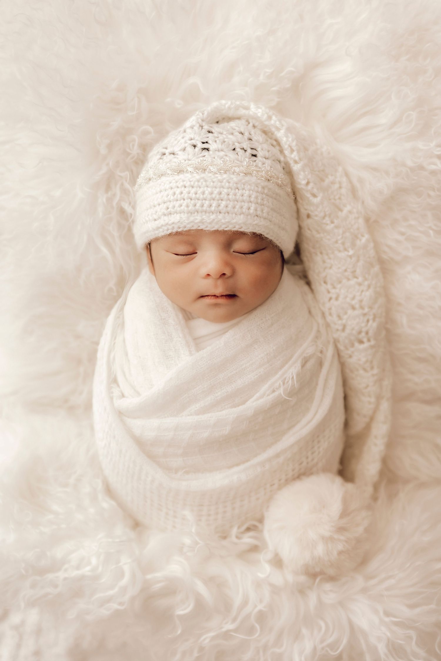 Sleeping newborn swaddled in soft white fabric and wearing a crochet hat rests on fluffy white fur backdrop. Professional newborn Photograph