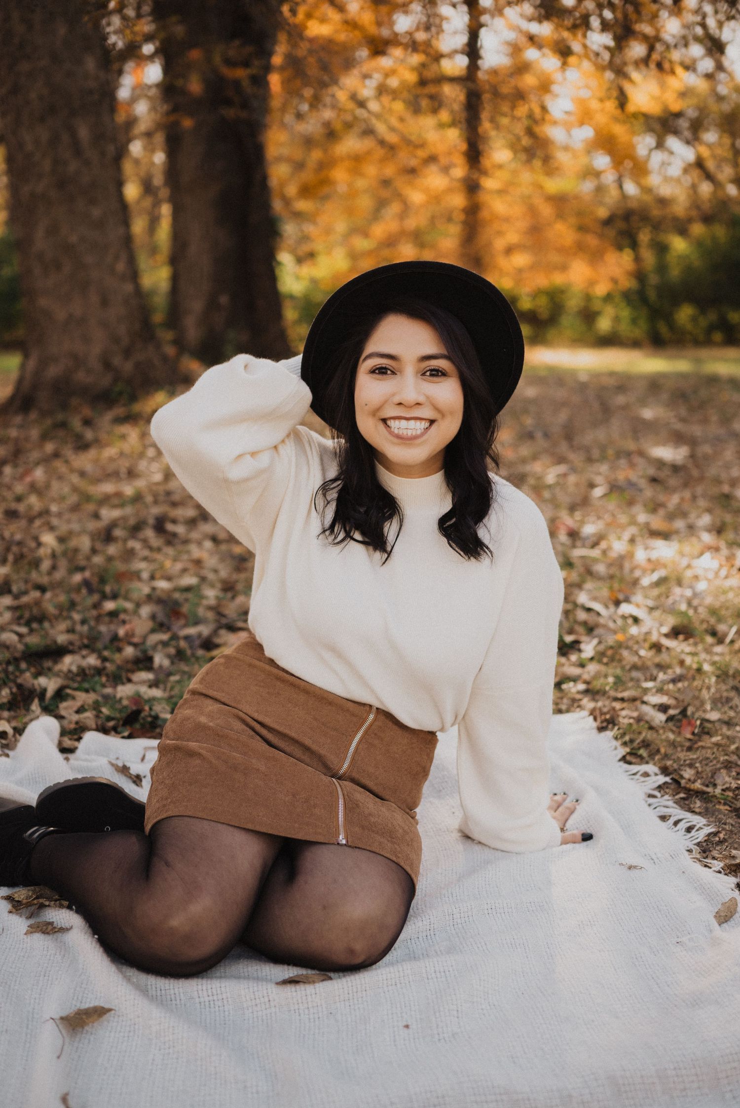 A person in a white sweater and brown boots lies on a blanket surrounded by autumn leaves.