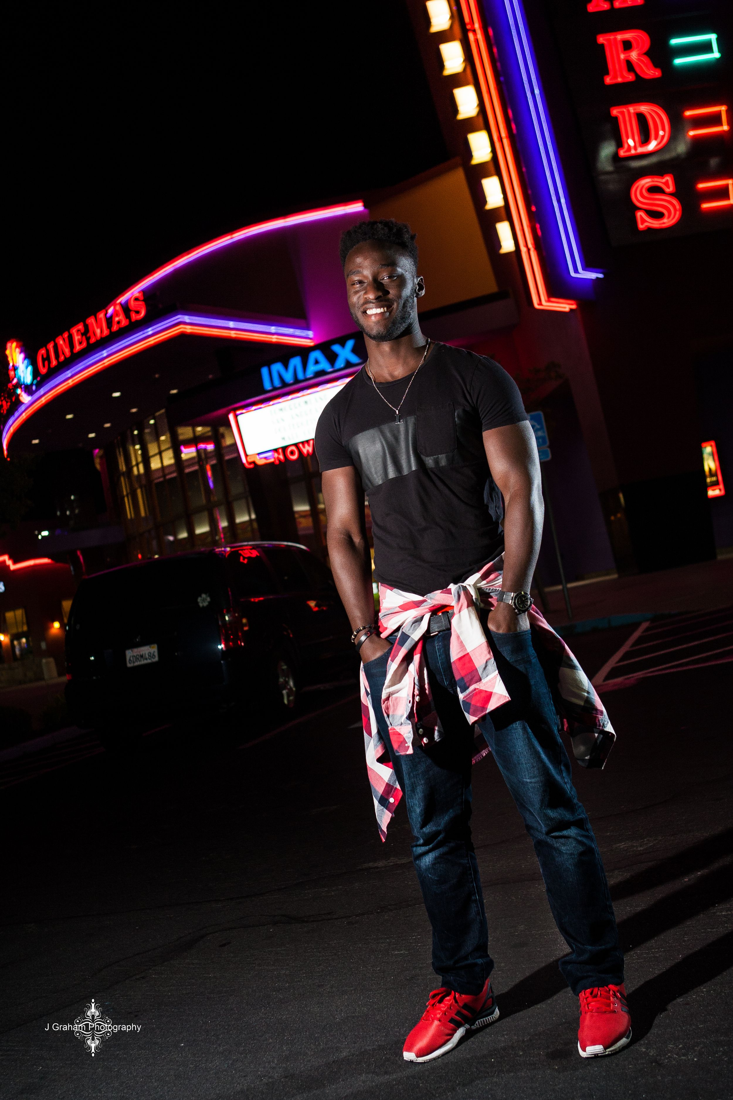 Person in black clothing and red sneakers poses in front of illuminated movie theater signs at night.