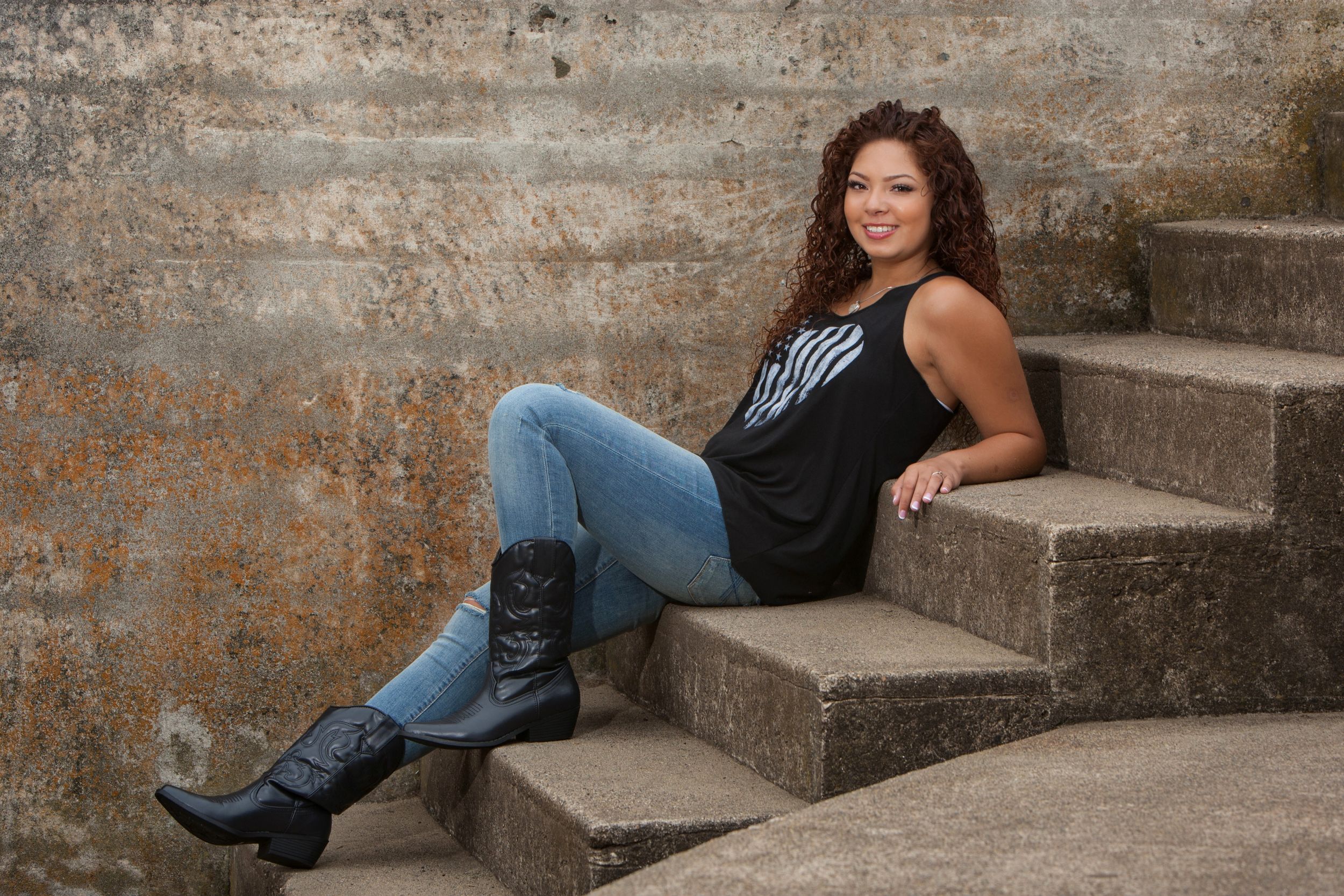 Woman in dark outfit poses on concrete steps against weathered brick wall background.