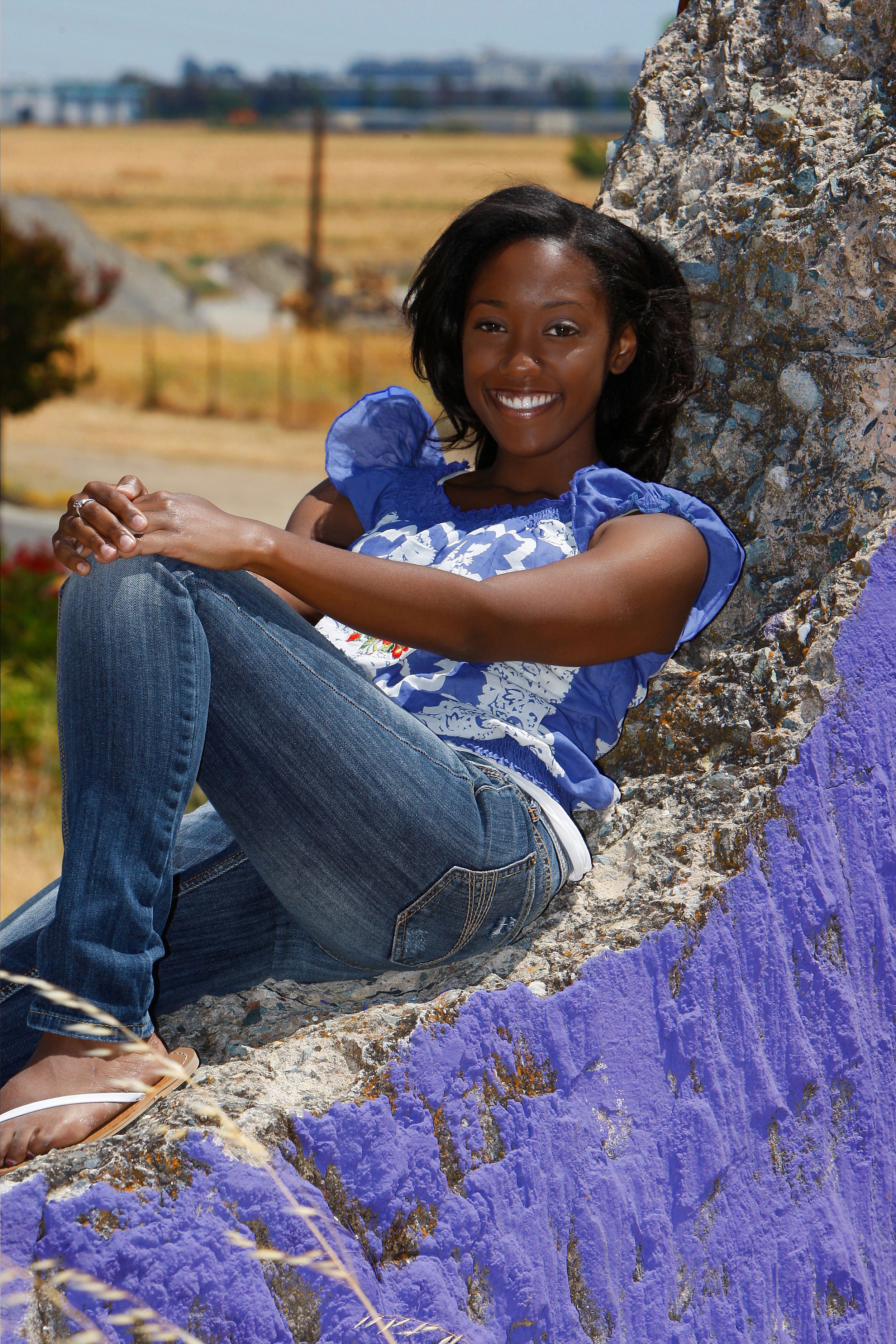 Person in blue top and jeans relaxes against a tree trunk surrounded by vibrant purple wildflowers in a field.