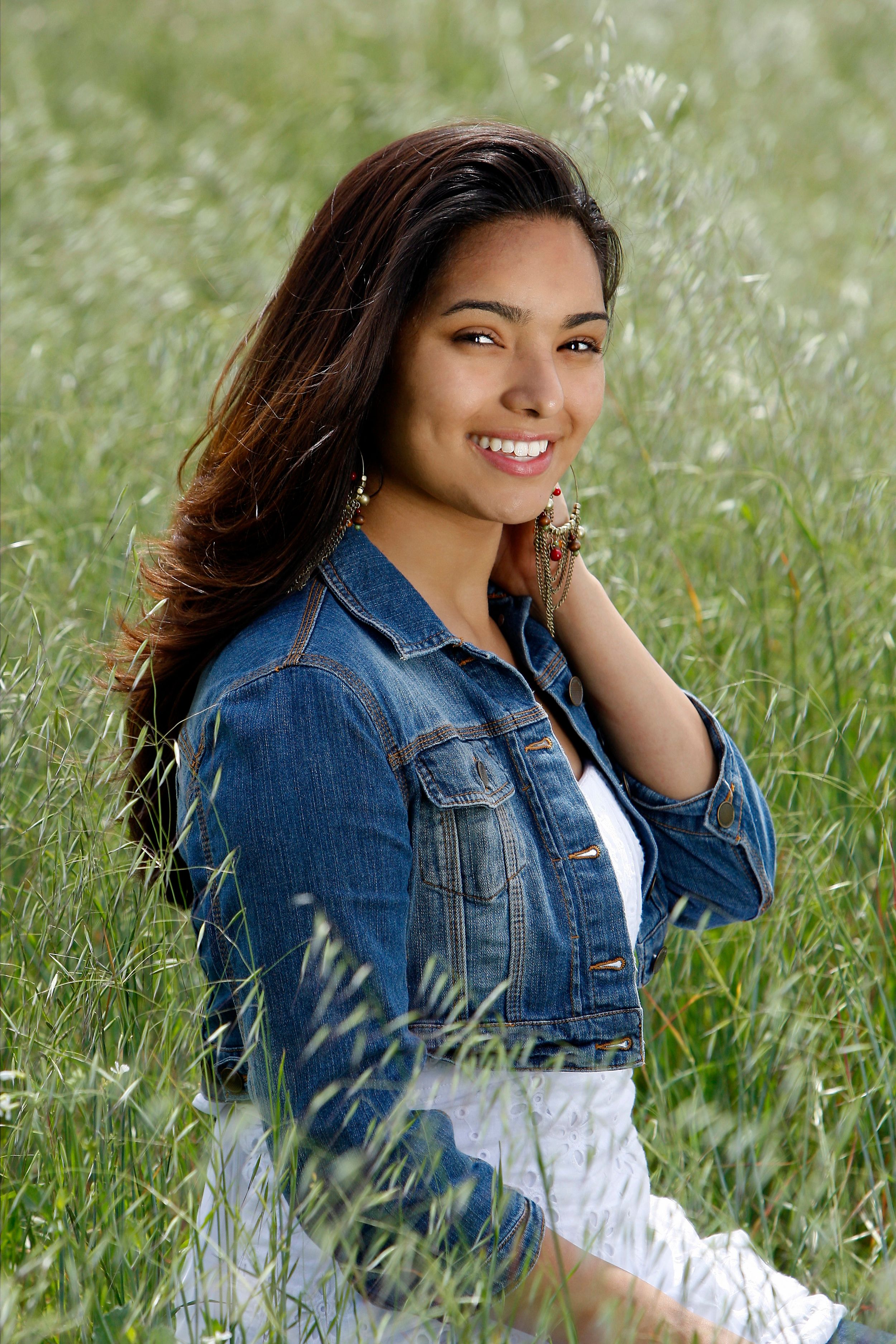 A person in a denim jacket poses outdoors in tall grass with a bright smile and natural lighting.