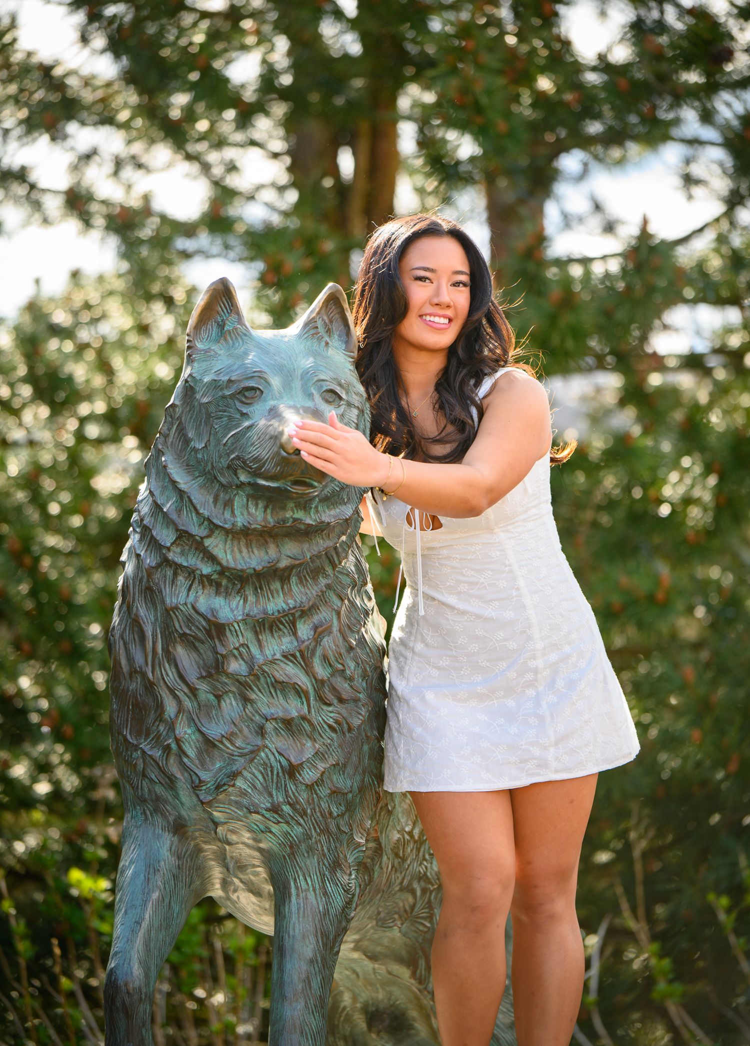 Graduate in white dress posing with UConn husky mascot statue outdoors.