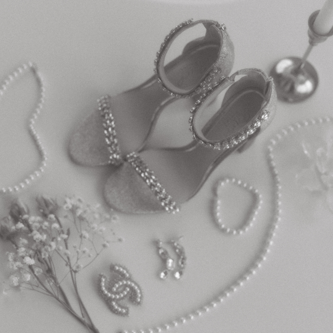 Wedding accessories arrangement with pearl necklace, silver shoes, and crystal details in monochrome.