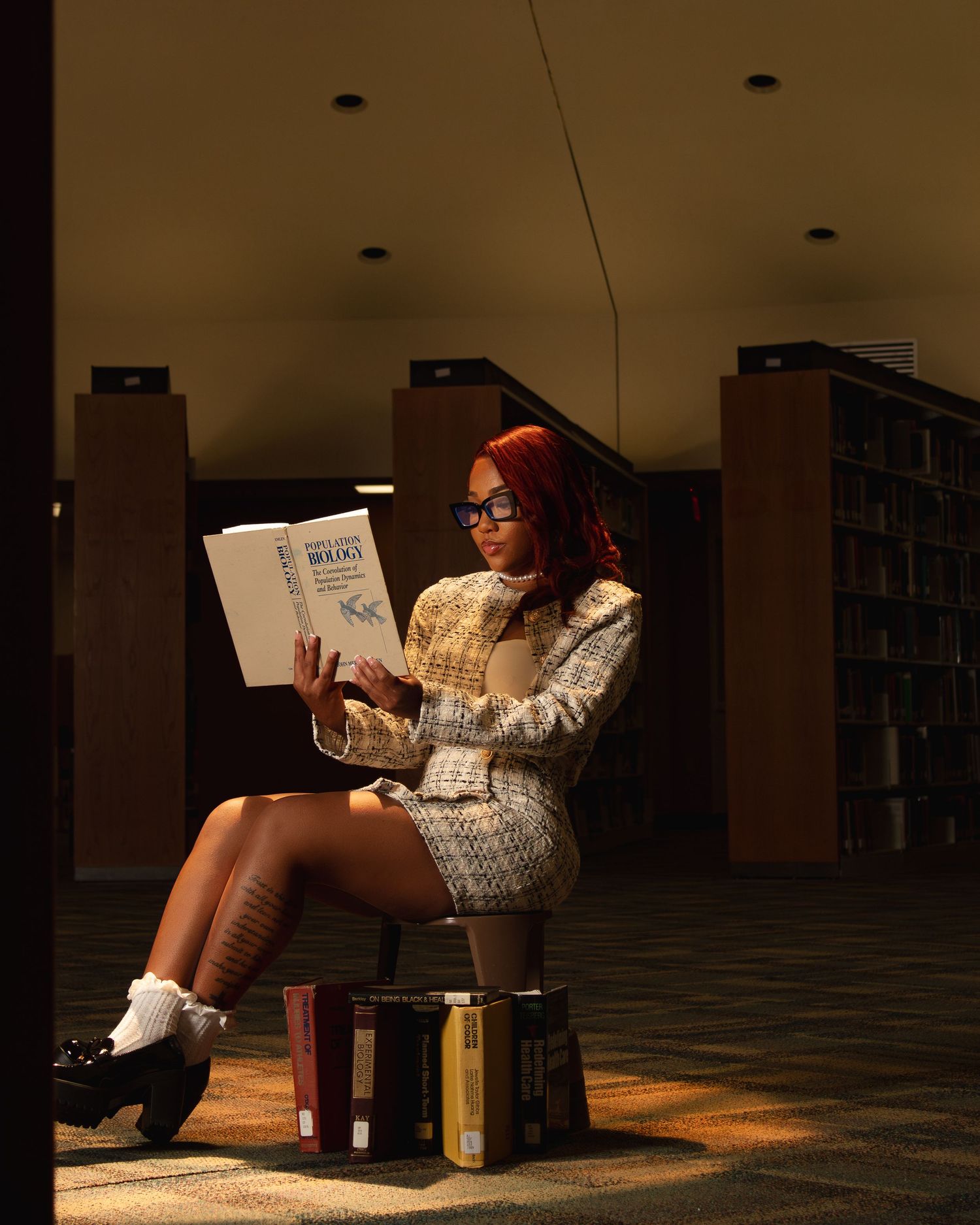 Student reading in library wearing cream sweater surrounded by stacked books.