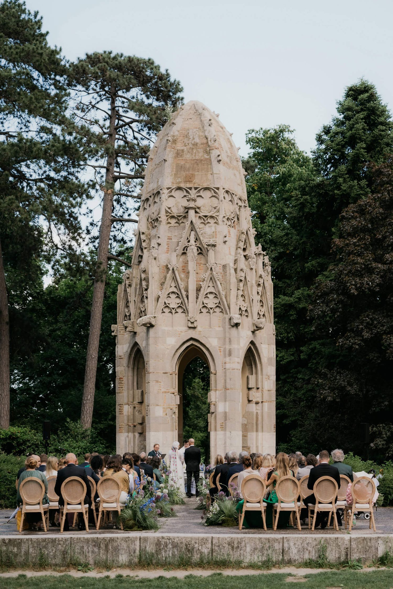 Outdoor wedding ceremony before ornate Gothic stone monument surrounded by lush trees and guests seated in rattan chairs.