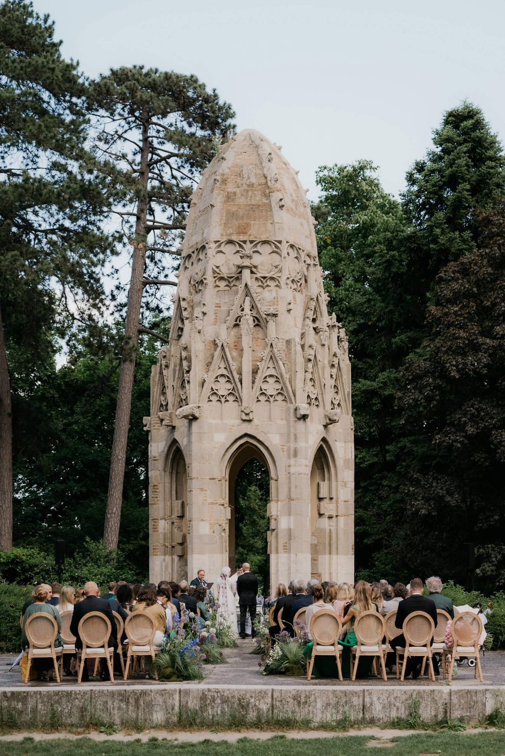 Outdoor wedding ceremony before ornate Gothic stone monument surrounded by lush trees and guests seated in rattan chairs.