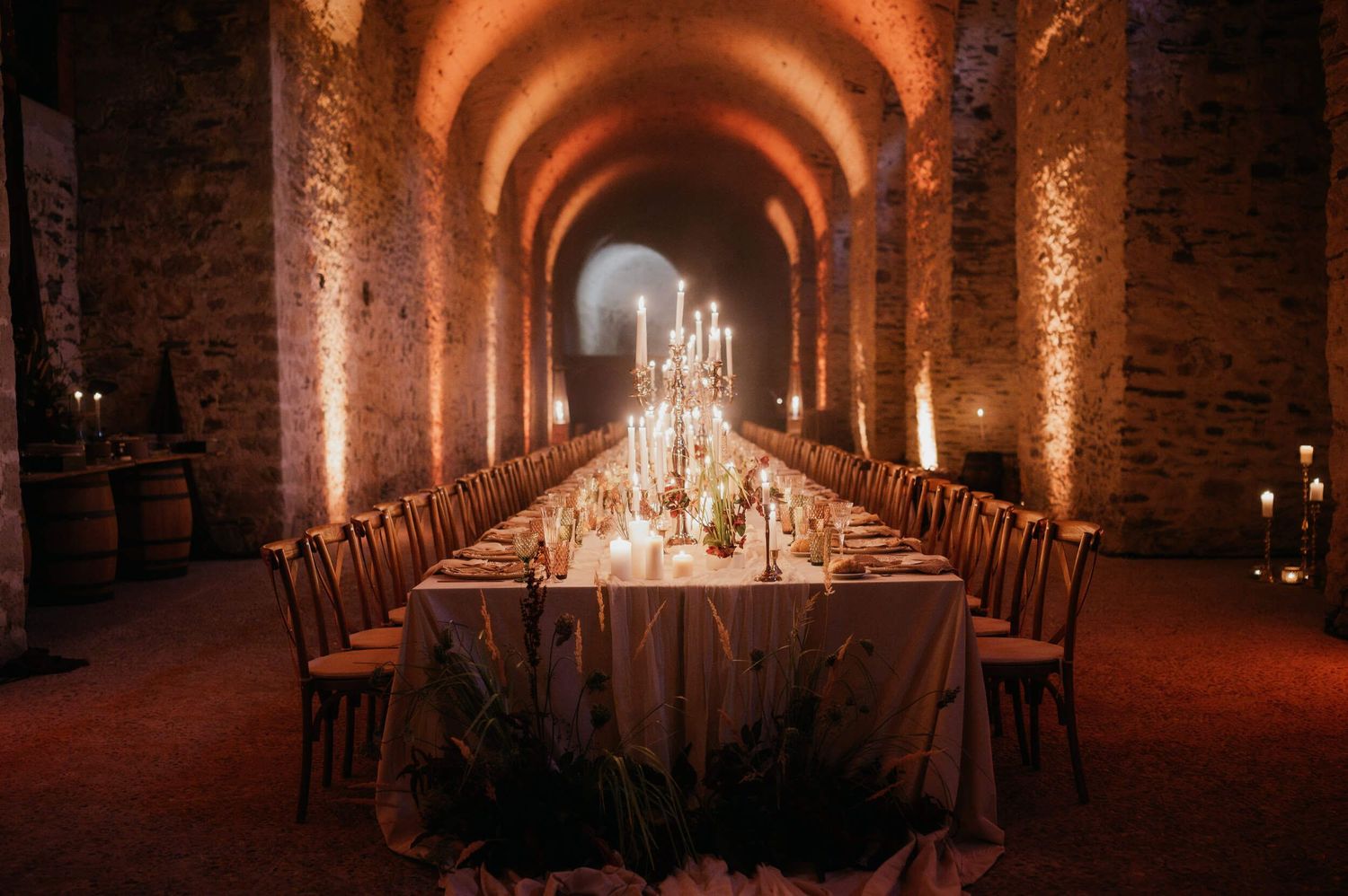 Long banquet table set for wedding reception in dramatic stone tunnel with candlelight.