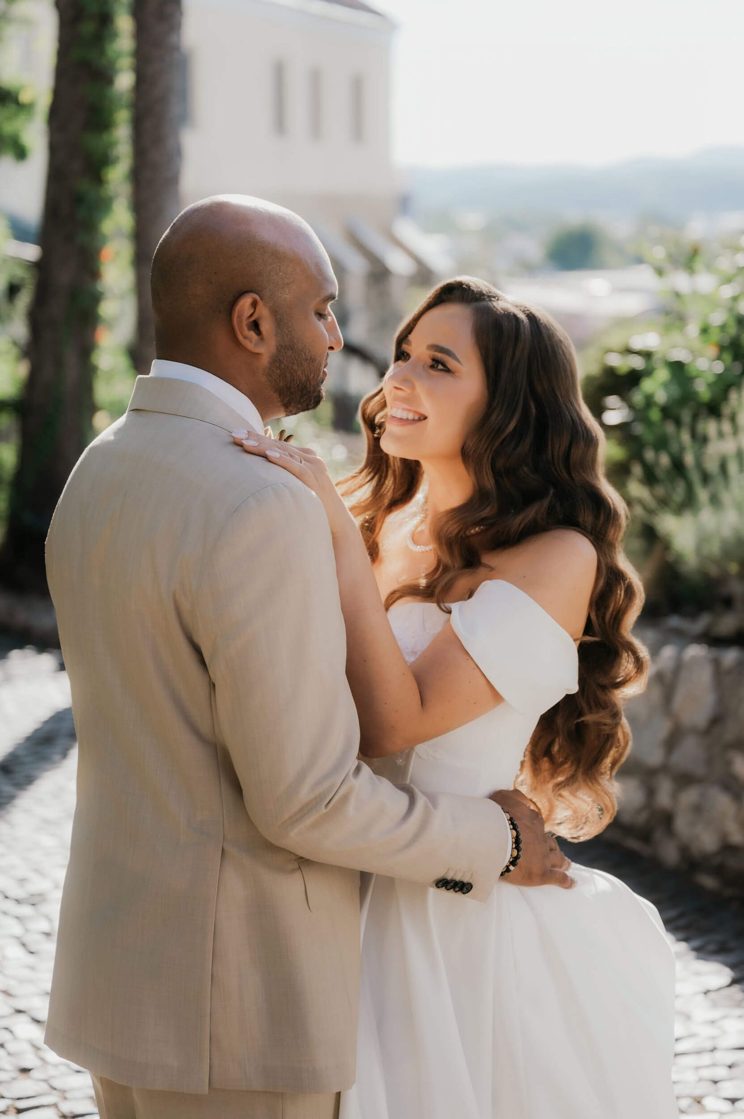 Couple in wedding attire embrace outdoors, bride in white off-shoulder gown smiling at groom in beige suit.