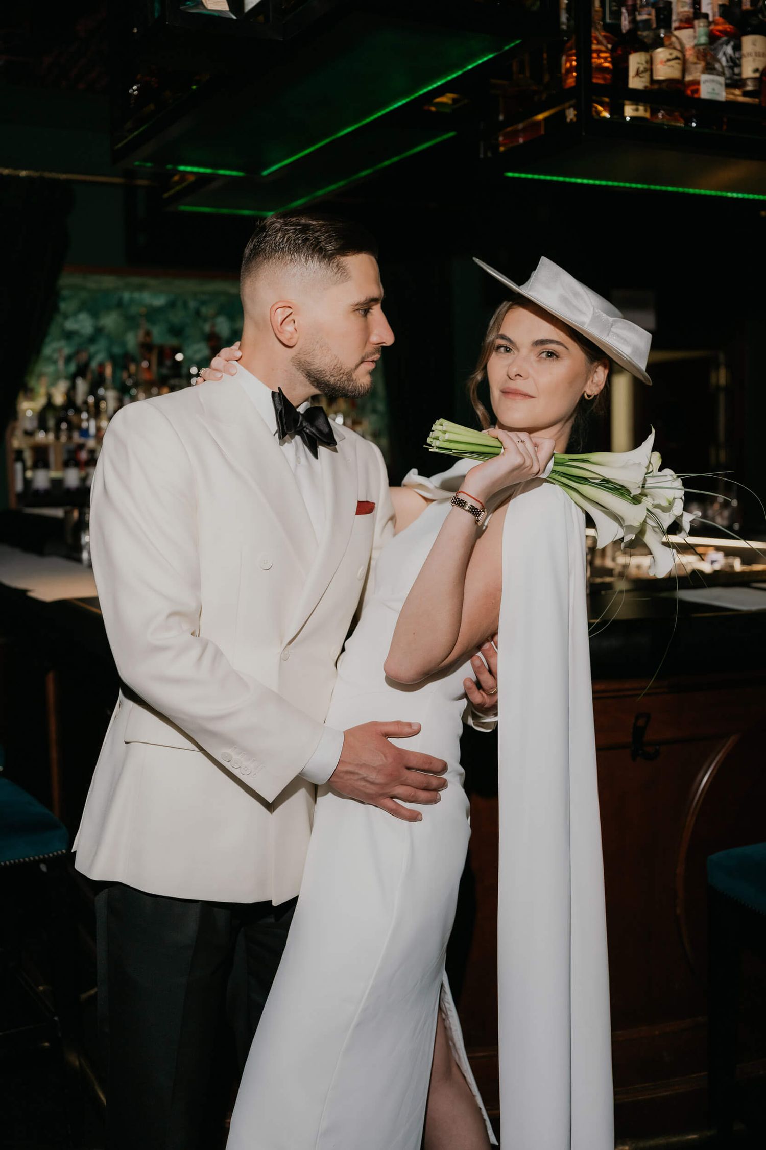 Elegant couple in white wedding attire pose at a bar, groom in tuxedo embracing bride in cape dress and hat.