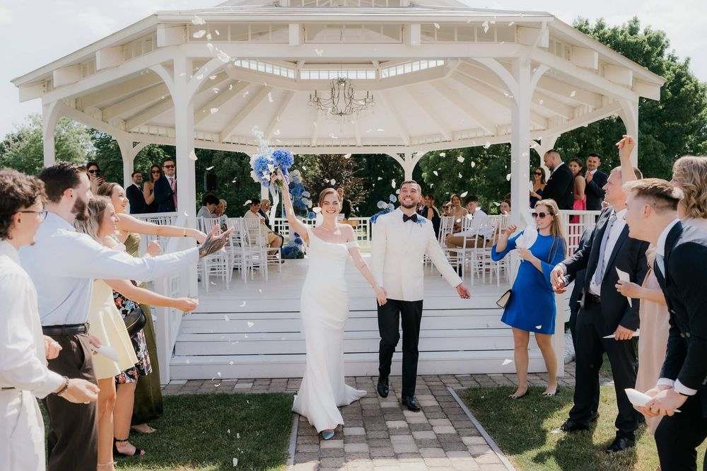 Bride and groom walk hand-in-hand through cheering guests outside a white gazebo at an outdoor wedding reception.