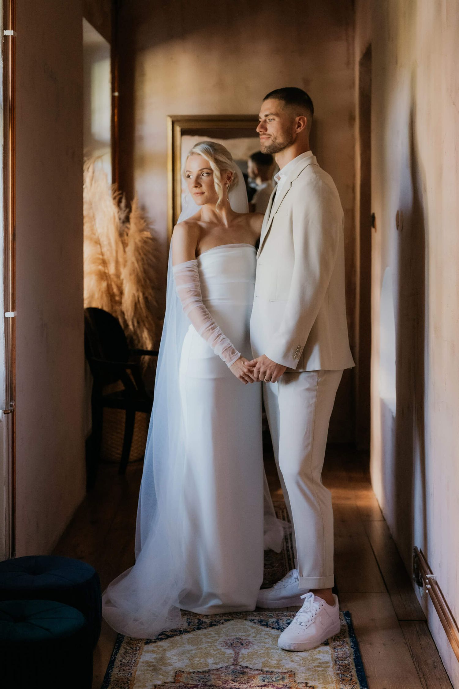 Bride and groom in white wedding attire hold hands in a warmly lit hallway with vintage rug.