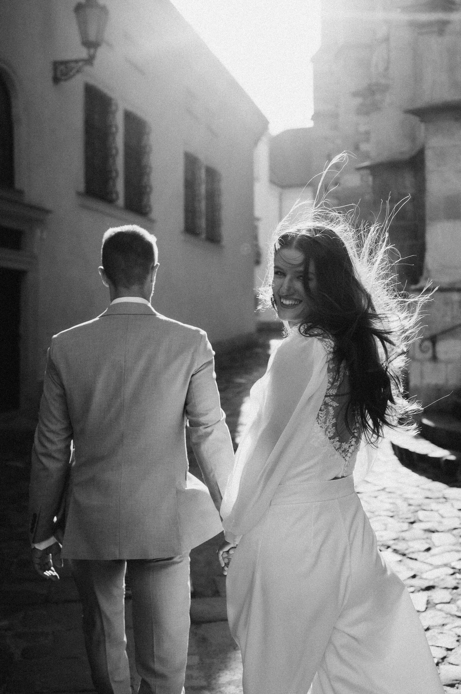 Black and white silhouette of couple in white dress and suit walking together in sunlit courtyard.