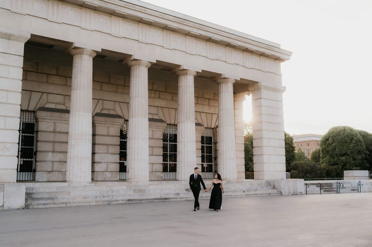 Couple in formal attire walking outside a grand neoclassical building with tall columns at sunset.