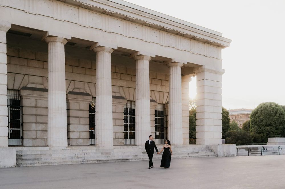 Couple in formal attire walking beside grand white columned building at sunset.
