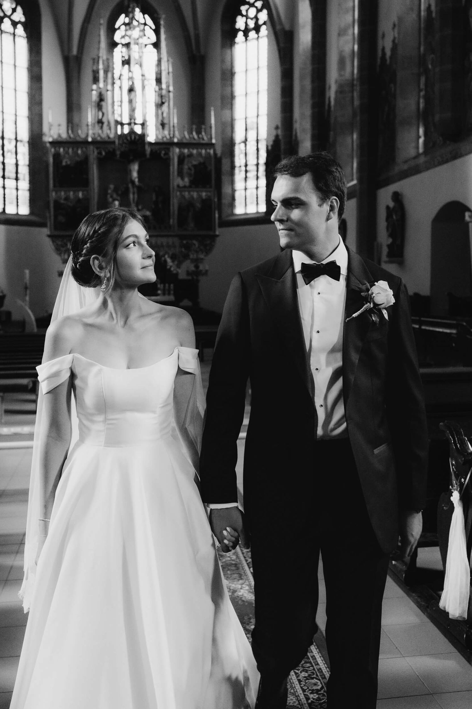 Black and white wedding photograph of couple in elegant attire walking inside a grand church with stained glass windows.