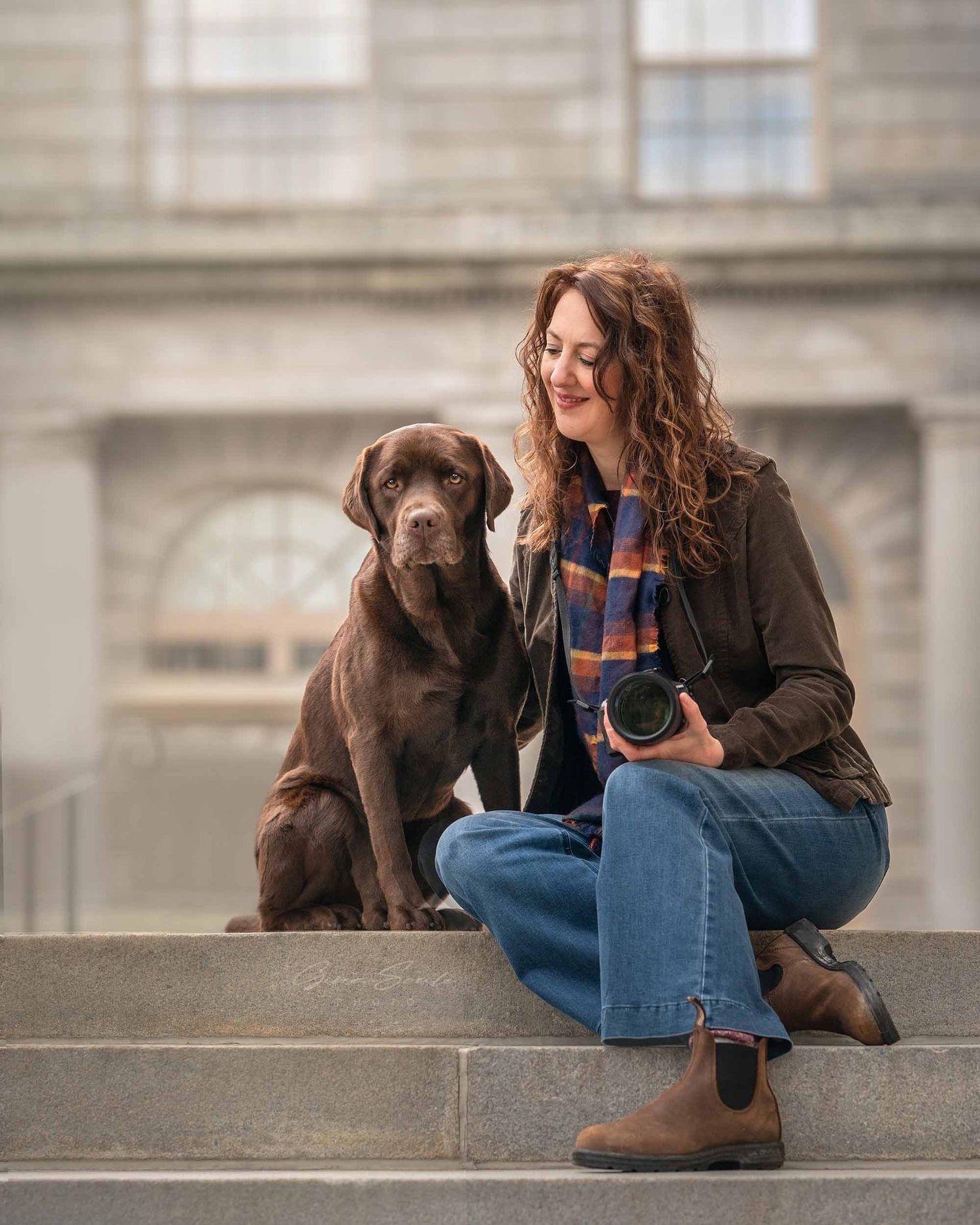 Jen with curly hair holding camera sits on steps beside her a chocolate Labrador. Xenophon Photography in Maine.