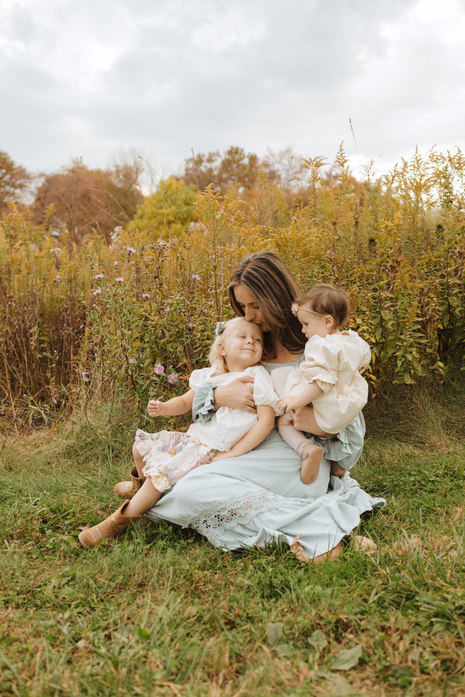 A mother and children sit together in a wildflower field on a cloudy autumn day wearing light colored clothing in tewksbury nj.