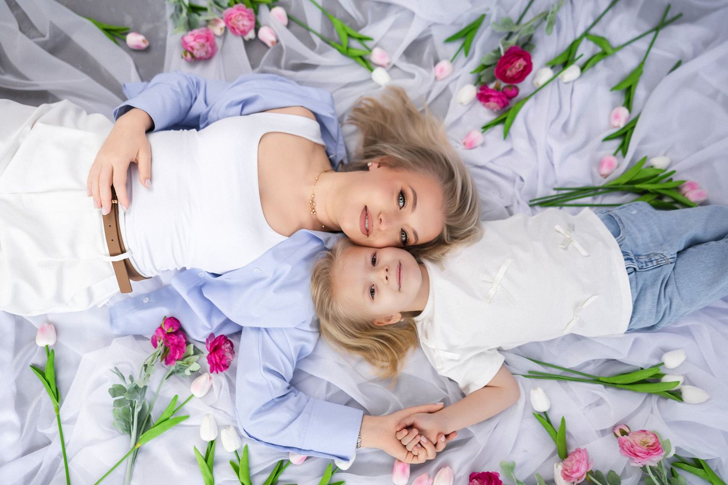 Mom and daughter lay on white fabric surrounded by pink and purple tulip flowers in a dreamy spring photo studio motherhood setup.