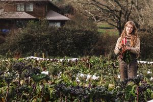 A gardener stands in a flourishing vegetable garden with a rustic house in the background during golden hour.