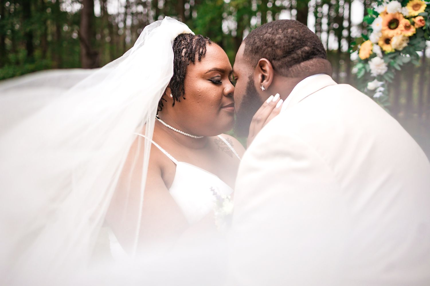 A Black bride and groom share a tender kiss outdoors, bride in white gown with flowing veil and sunflower backdrop.