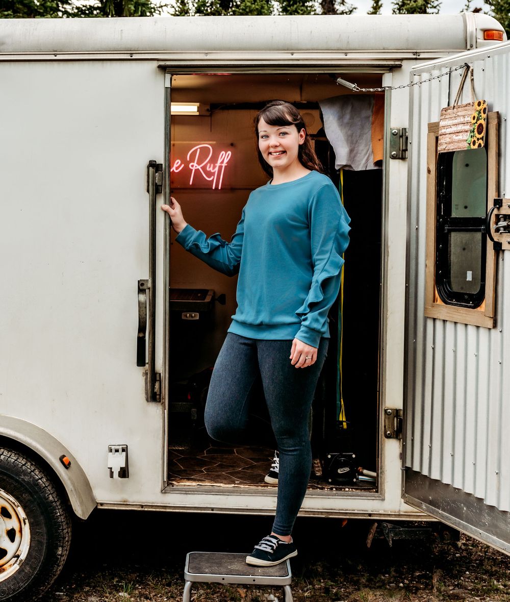 A person poses cheerfully in the doorway of a mobile food trailer with neon signage visible inside.