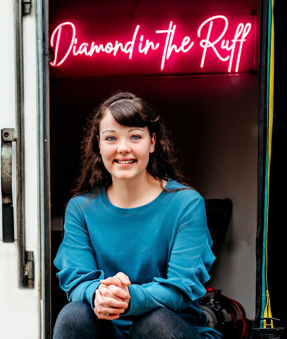 A bright neon sign reading 'Diamond in the Ruff' glows above someone in a teal sweater.
